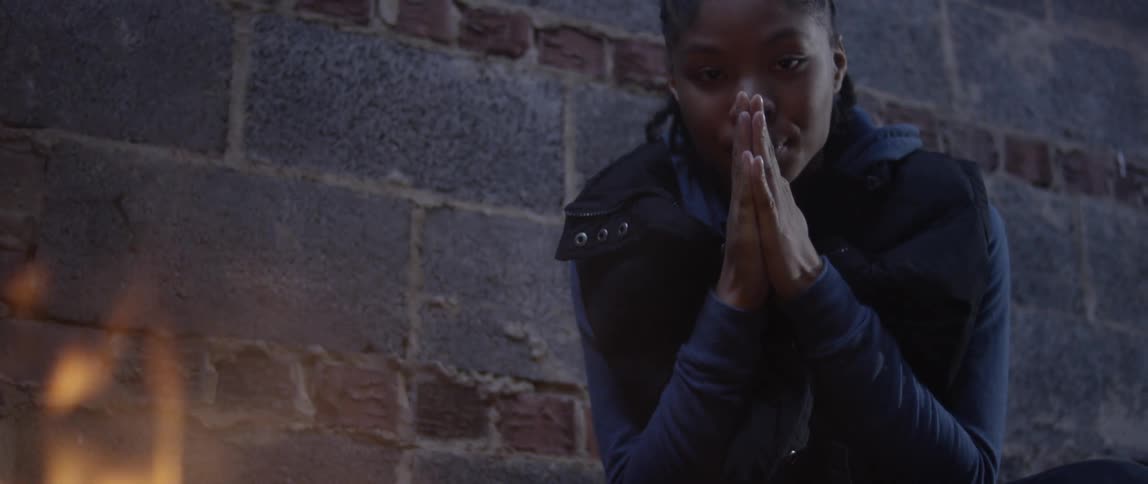 A young black woman warms her hands at a fire with a brick wall in the background