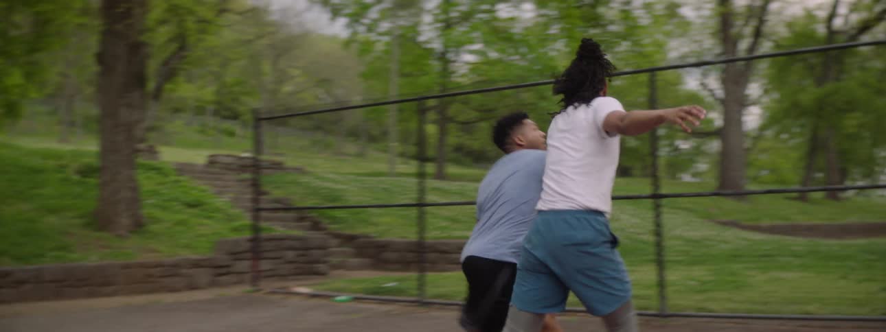 A young man skillfully dribbles past a defender and makes a layup on an outdoor basketball court in slow motion