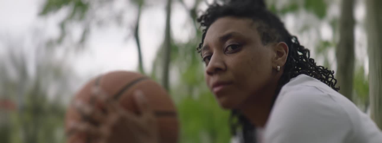 A determined female basketball player sits with her ball in the park and gives an intense look to the camera