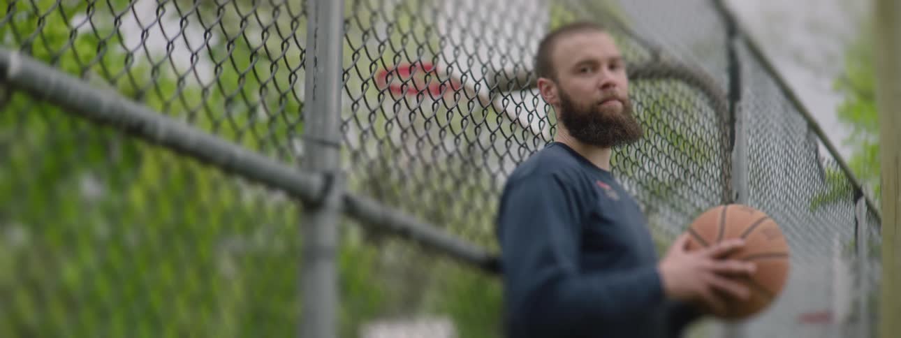 A chain-link fence is in focus as a pensive, bearded man holding a basketball stands behind it on an outdoor court