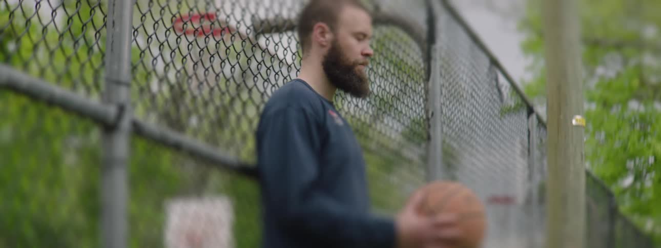 A focused bearded man stands by a chain-link fence on an outdoor basketball court, contemplating his next move in slow motion