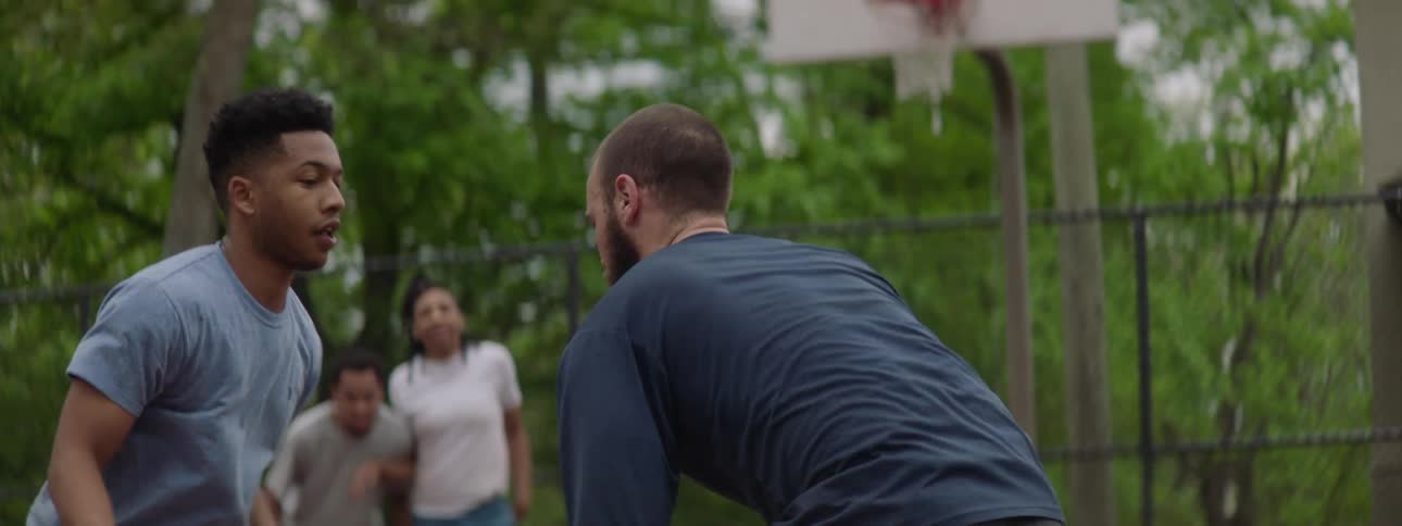 A bearded man dribbles a basketball against a defender in slow motion on an outdoor court