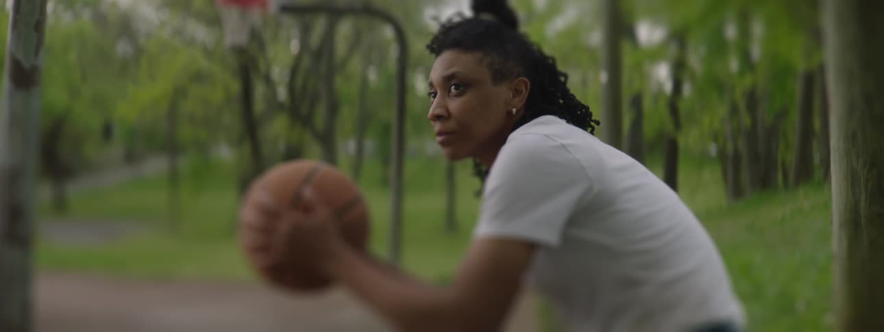 A female basketball player with a focused expression sits on an outdoor court holding a ball in slow motion