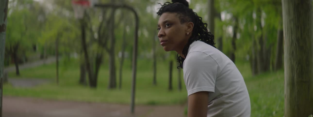 A determined basketball player sits contemplatively on the side of an outdoor court in slow motion