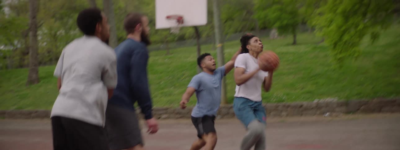 A woman skillfully dribbles past her friends and scores a layup in slow motion during a basketball game at an outdoor park