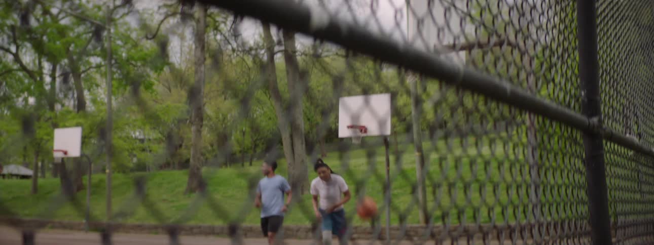 A man makes a clean shot during a friendly game of basketball at an outdoor park court, viewed through a fence in slow motion