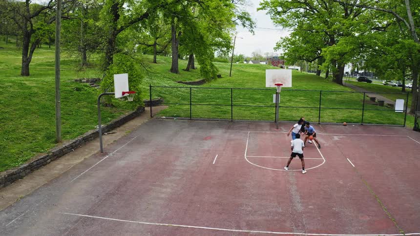 A high-angle aerial view captures a group of friends playing a friendly game of pickup basketball on an outdoor court surrounded by a lush green park