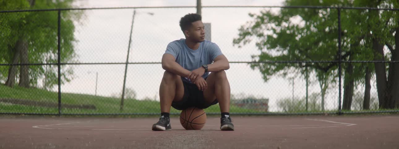 An athletic young man takes a thoughtful break, sitting on his basketball on an outdoor court before looking into the camera