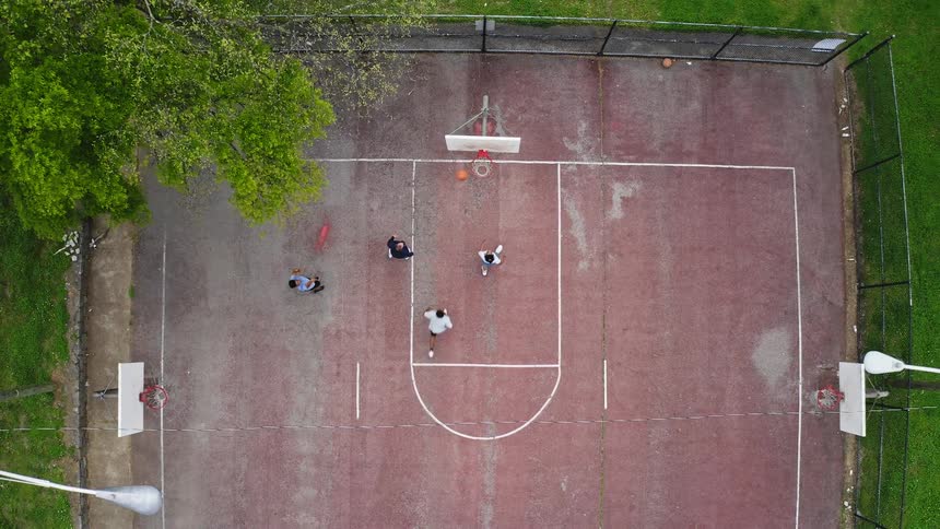 A rising top-down drone shot captures three friends enjoying a casual game of basketball on an outdoor court