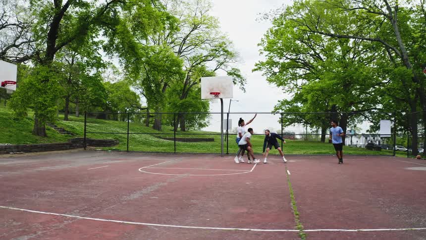 A diverse group of friends enjoy a slow-motion game of pickup basketball on an outdoor court in a park