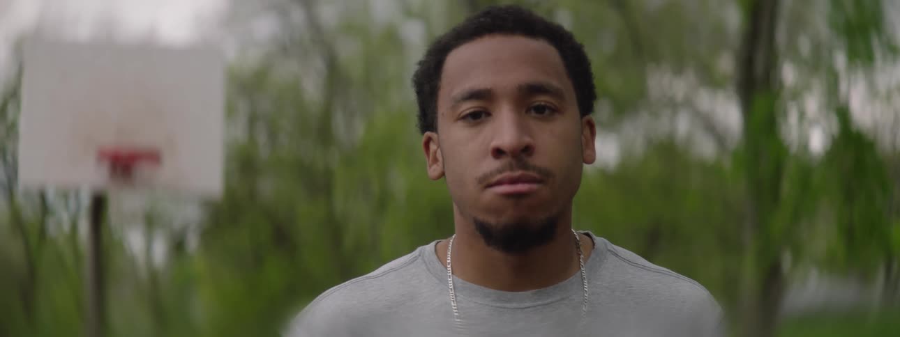 A young Black man with a serious expression slowly turns to look at the camera on an outdoor basketball court