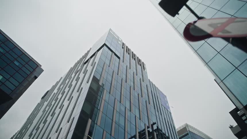 A low-angle shot pulls back from a traffic sign to reveal the impressive facade of modern glass skyscrapers under a grey, overcast sky