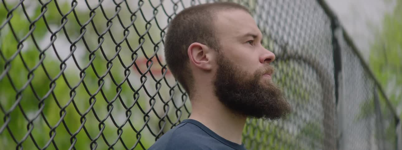 A thoughtful bearded man rests against a chain-link fence on a basketball court as the camera focus shifts from the hoop in the background to him