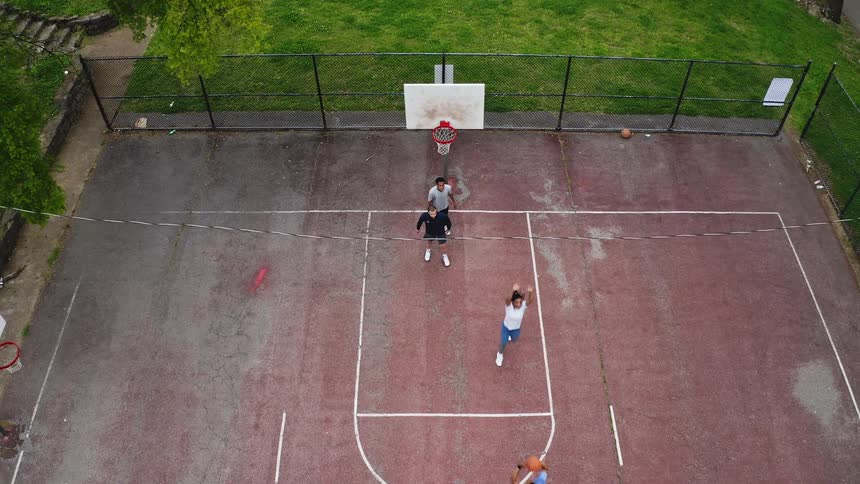 A slow-motion, high-angle aerial shot descends over a group of friends playing a casual basketball game on an urban park court