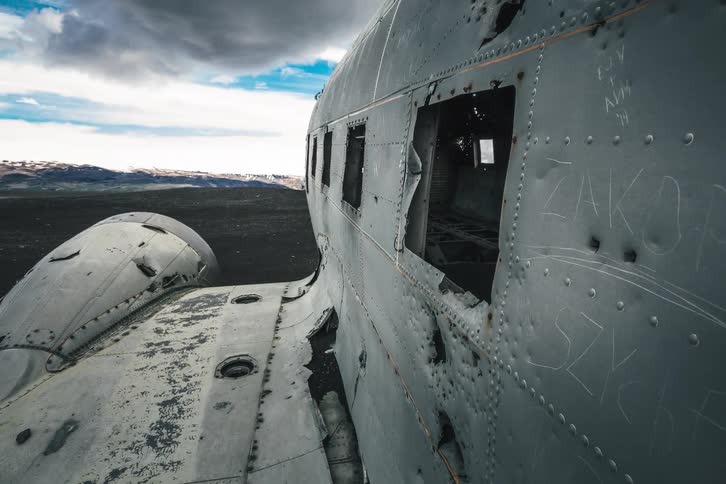 Eerie tracking shot from the dark, sand-filled interior of a crashed plane to its desolate, weathered exterior on a black volcanic plain