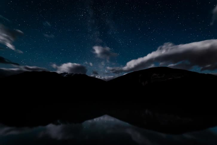 A serene time-lapse captures the Milky Way and stars rotating above a mountain lake, with their reflections shimmering on the water as clouds sweep across the night sky