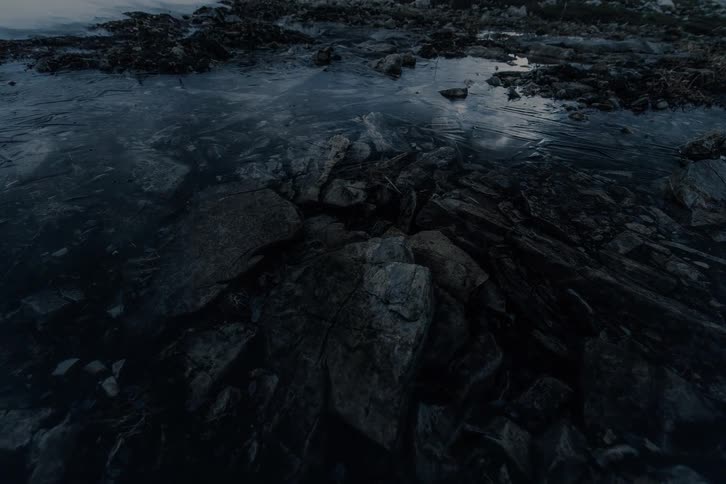 A close-up, slow pan reveals dark, jagged rocks emerging from a layer of melting ice in a cold, desolate landscape