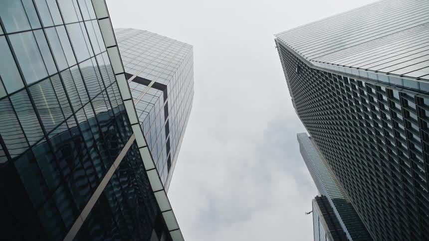 A slow, low-angle shot captures the impressive scale of modern glass skyscrapers against an overcast sky