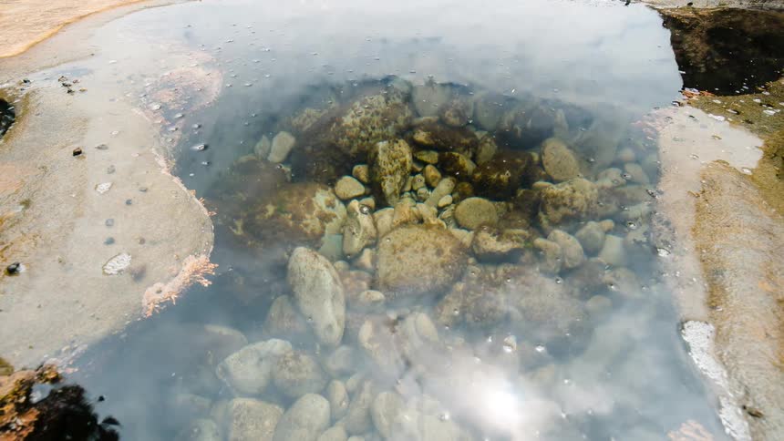 Time-lapse of a rocky tide pool where the water's surface reflects passing clouds and the sun as the camera zooms out
