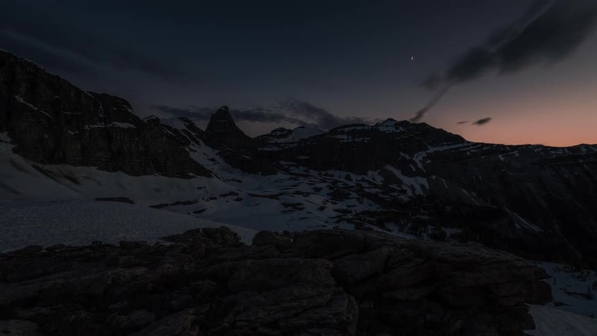 A quiet dusk settles over a snow-covered mountain range as clouds drift by and a crescent moon appears in the sky