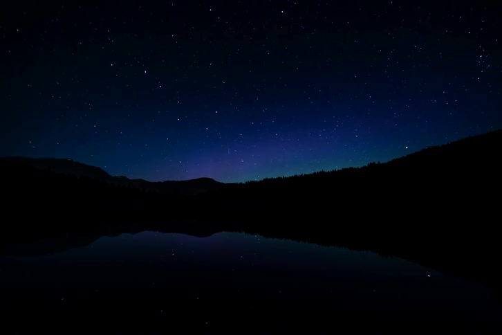 A vibrant purple and green Aurora Borealis dances across a star-filled night sky, casting a beautiful reflection on a tranquil mountain lake