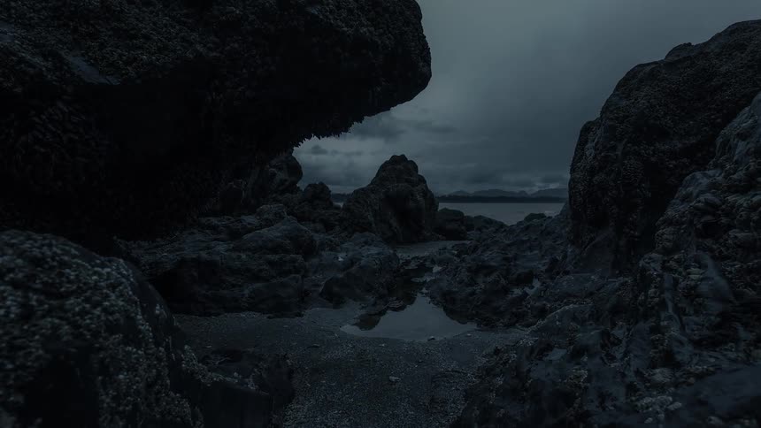 A slow dolly shot moves through a dark, rocky cove towards the ocean under a gloomy, overcast sky