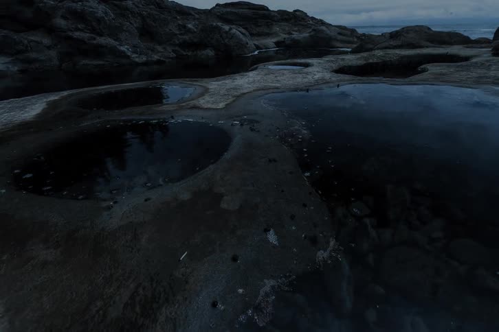 A slow pull-back reveals dark, still tide pools dotting a rocky coastline under a moody, overcast sky