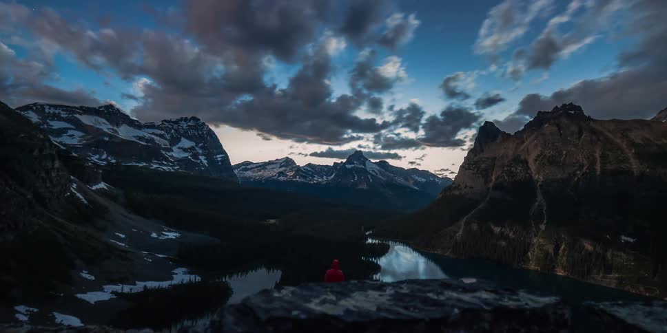 A breathtaking time-lapse captures a hiker in a red jacket standing victoriously on a cliff, overlooking a pristine mountain lake and valley as morning clouds roll by