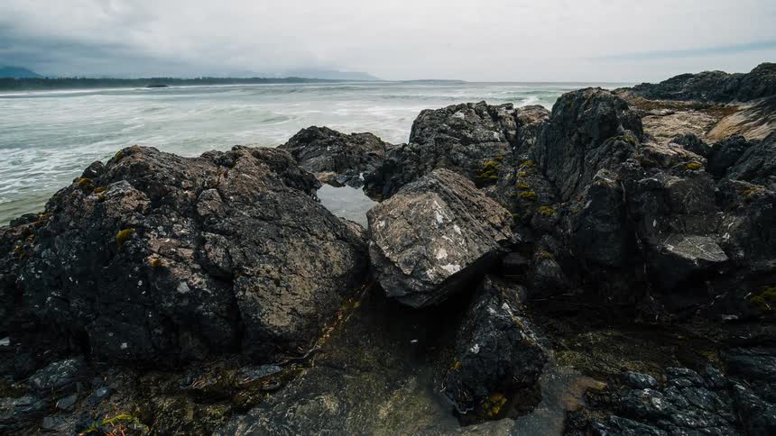 Slowly tilting up from a tide pool among rugged rocks to reveal the expansive and turbulent ocean on an overcast day