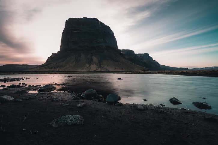 A majestic, dark mountain is silhouetted against a colorful sunrise as a river flows gently by on a rocky, dark sand shore in this slow push-in shot