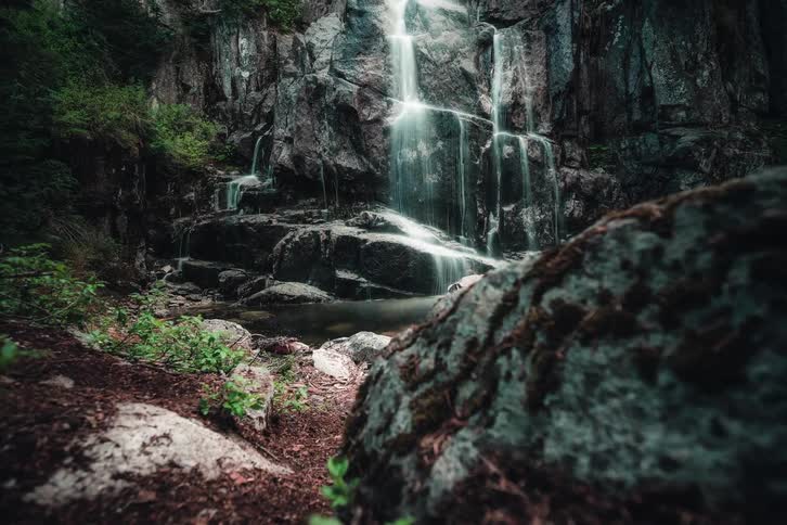 A dramatic timelapse pulls back to reveal a beautiful waterfall cascading over dark, mossy rocks in a lush forest