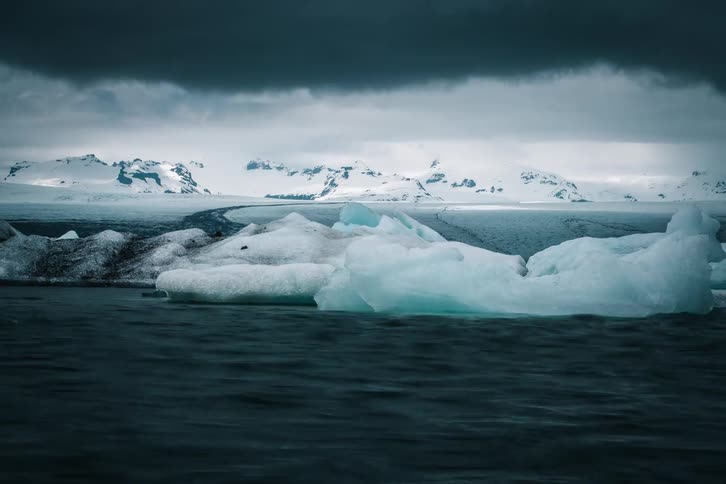 A dramatic timelapse shows large icebergs floating across a dark glacial lagoon in front of a vast, snowy mountain range