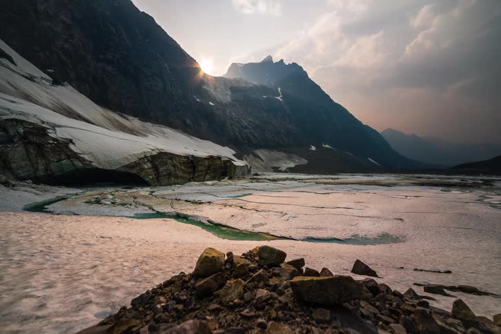 The sun sets behind a rugged mountain peak in a stunning timelapse, illuminating a melting glacier and casting long shadows across the icy landscape
