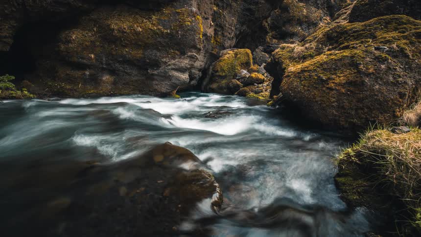 A stunning long exposure time-lapse captures the silky smooth water of a river flowing through a dramatic, moss-covered rock canyon