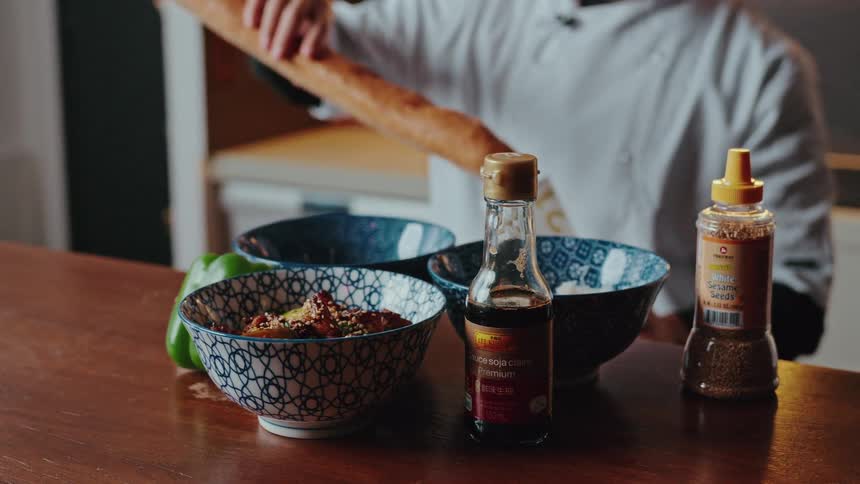 A close-up of a prepared Asian dish and ingredients like soy sauce and sesame seeds on a counter, with a chef working in the background