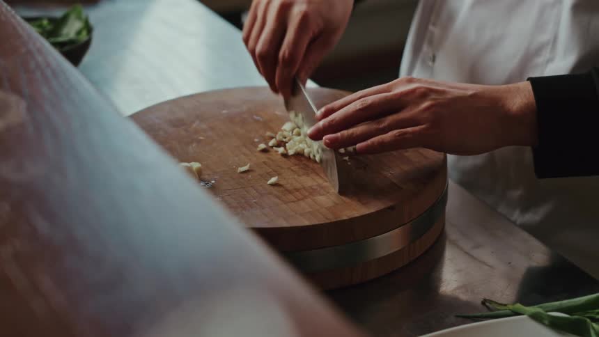 A professional chef in a kitchen meticulously minces fresh garlic with a cleaver on a round wooden cutting board
