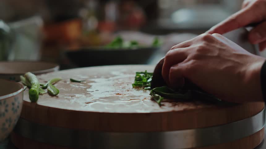 A chef's hands expertly chop fresh green onions with a cleaver on a wooden cutting board in a slow-motion close-up
