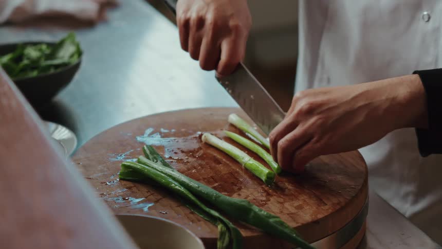 Close up of a professional chef's hands skillfully chopping fresh green onions on a wooden board in a restaurant kitchen