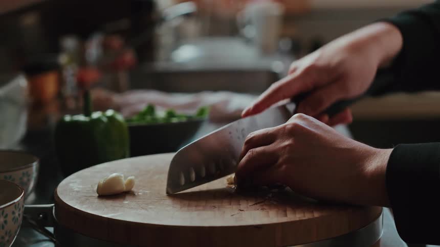A person in slow motion minces garlic with a sharp knife on a round wooden cutting board in a cozy kitchen