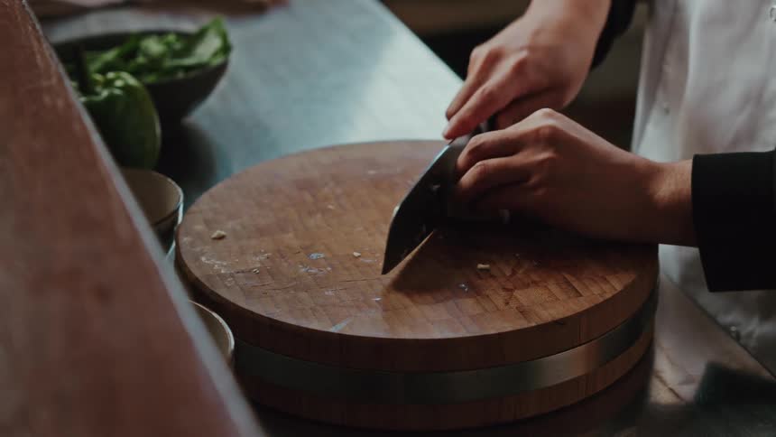 A professional chef crushes and then minces garlic on a circular wooden cutting board in a restaurant kitchen