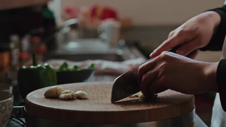 Slow-motion close-up of a professional chef's hands skillfully mincing a clove of garlic on a thick wooden cutting board
