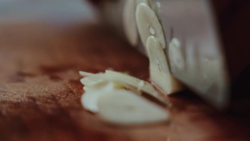 Extreme close up of a sharp knife slicing a garlic clove into thin pieces in slow motion on a wooden board