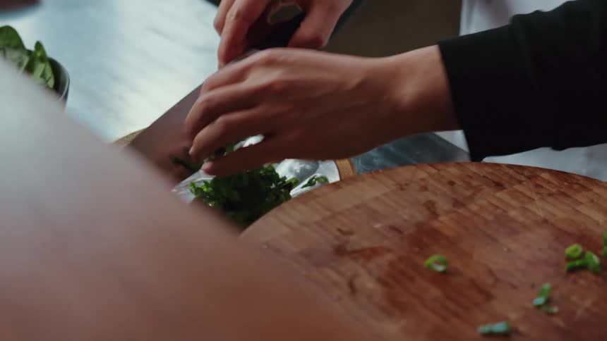 A chef carefully slides freshly chopped green onions from a wooden cutting board onto a glass plate in slow motion