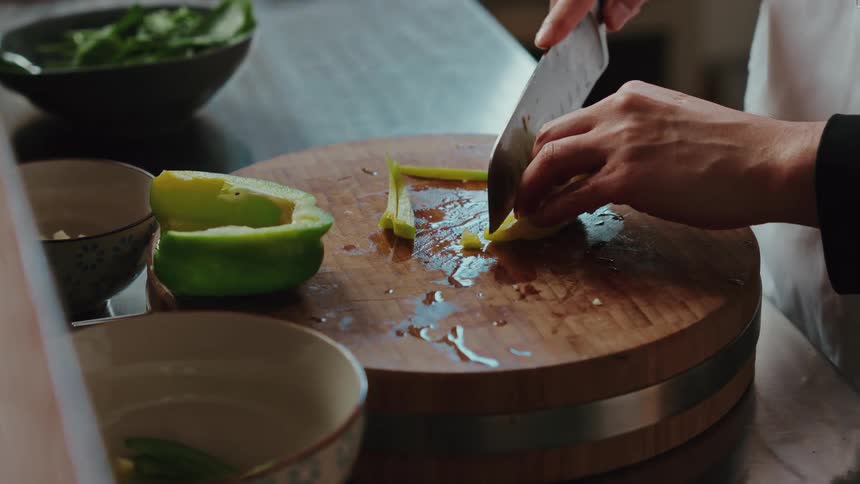Slow motion close up of a chef expertly slicing a green bell pepper on a wooden cutting board in a professional kitchen
