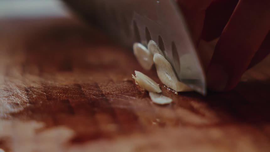 A close-up, slow-motion shot of a person slicing a garlic clove on a wooden cutting board with a sharp knife
