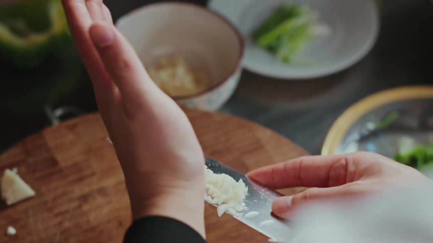 Close-up, over-the-shoulder view of a chef mincing garlic on a wooden cutting board in slow motion