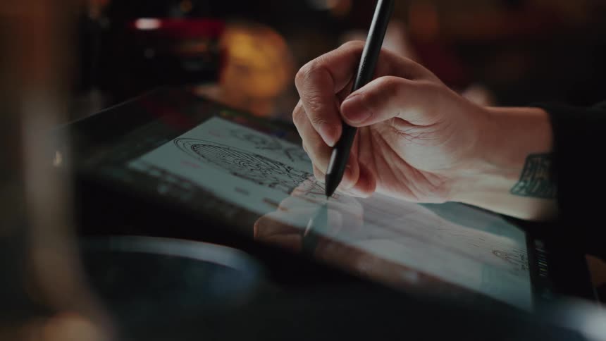 An artist with a tattooed wrist uses a stylus to create a detailed digital drawing on a graphics tablet in a dimly lit studio