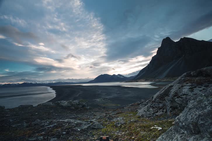 A dramatic pull-back time-lapse showcases a vast, rugged landscape with a dark sand beach and majestic mountains under a moody, cloud-filled sky