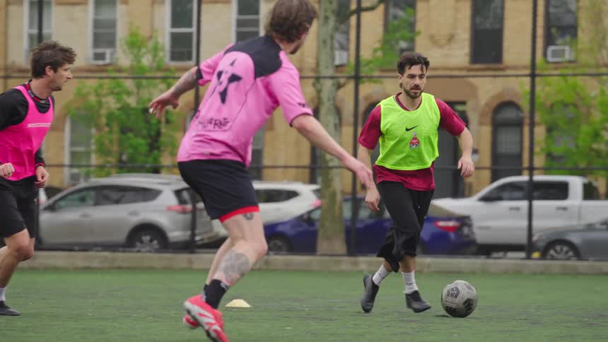 A male soccer player in a yellow vest expertly dribbles the ball past a defender before making a pass in slow motion on a city field