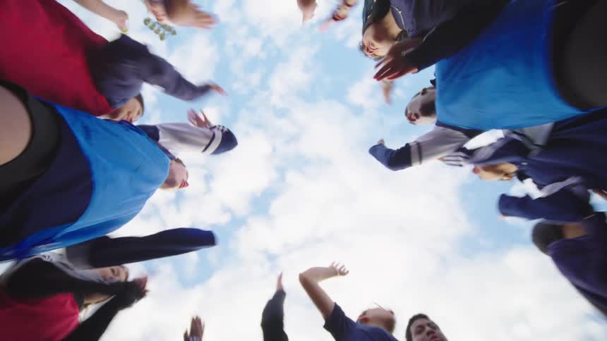 A low-angle view of a diverse sports team huddling together, putting their hands in for a cheer before breaking apart with energy under a blue sky